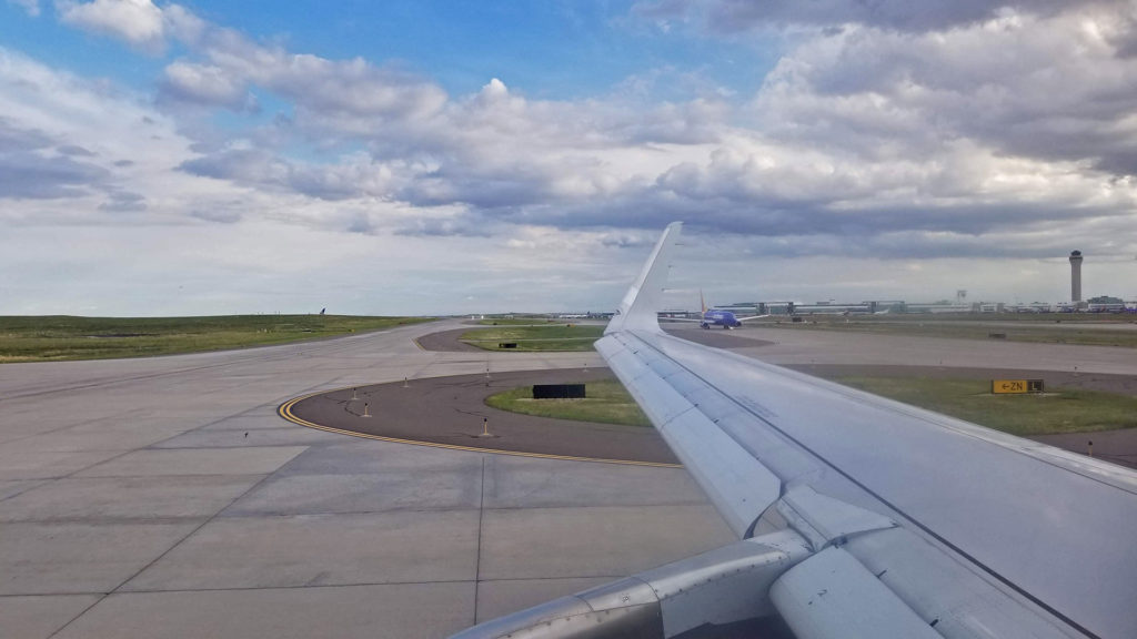 Airplane wing with runway and clouds in the background.